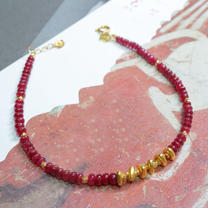 May include: A delicate bracelet featuring small, round, deep red beads interspersed with gold-colored accents. The bracelet has a gold-colored clasp and a small chain for adjustable sizing. The design is simple yet elegant, suitable for everyday wear.