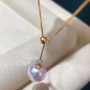 Puede incluir: Un delicado collar de oro con un colgante de perla iridiscente. La perla es de color lavanda suave con sutiles reflejos azules y rosas. El collar tiene una cadena fina y una cuenta deslizante para una longitud ajustable, mostrando un diseño minimalista.