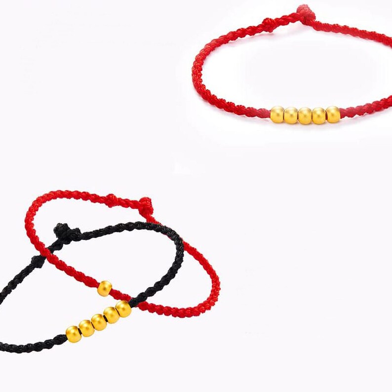 May include: Red and black braided bracelets with gold-colored beads. One bracelet is red with five beads, and the other is black with four beads. The bracelets are displayed against a white background.