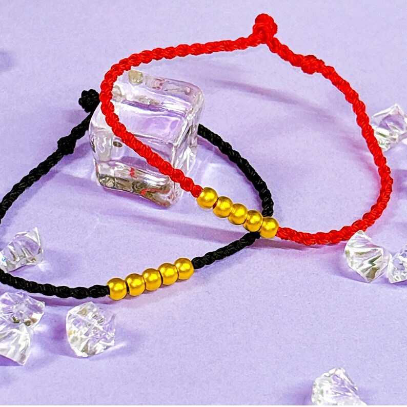 May include: Two adjustable bracelets. One is red, the other is black. Each bracelet features a series of gold-colored beads. The bracelets are displayed on a purple surface with clear, faceted decorative accents.