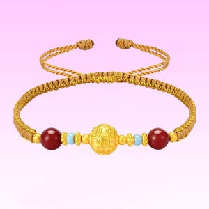 Peut inclure: Bracelet doré avec une bande tressée et des liens réglables. Le bracelet présente une sphère centrale dorée avec un motif en relief, flanquée de deux perles rouges et de petites perles bleues. Le fond est un dégradé de rose et de blanc.