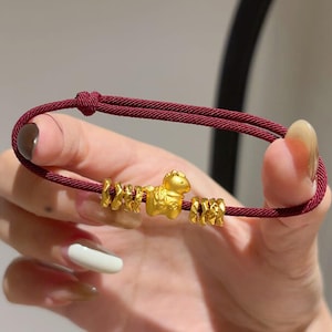 May include: A burgundy cord bracelet with gold-coloured beads and a horse charm. The bracelet is held in a hand, highlighting the design and the knot closure. The horse charm is the central feature of the bracelet.