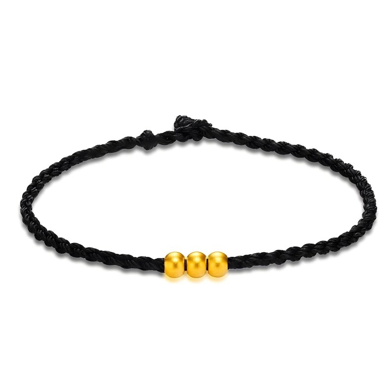 May include: A black braided bracelet featuring three gold-colored beads. The bracelet is made of a dark, woven material and has a simple, elegant design. The beads are evenly spaced and add a touch of shine.