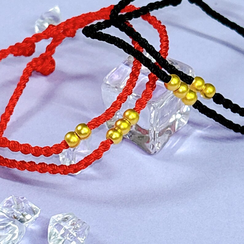 May include: Close-up of red and black braided bracelets with gold-colored beads. The bracelets are displayed on a reflective surface with clear, faceted objects in the background. The bracelets are likely for fashion or spiritual purposes.