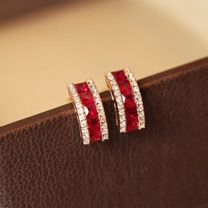 Peut inclure: Une paire de boucles d'oreilles créoles dorées. Chaque boucle d'oreille présente une rangée de pierres précieuses rouges vibrantes, encadrée par une bordure de petites pierres blanches scintillantes. Les boucles d'oreilles sont posées sur un fond marron.