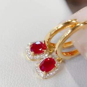 Puede incluir: Pendientes de aro dorados con un colgante de piedra preciosa roja ovalada, rodeado de pequeñas piedras claras. Los pendientes se muestran sobre una superficie clara.