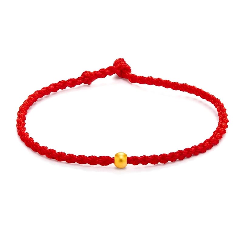 May include: A red braided bracelet with a single gold bead. The bracelet is made of a tightly woven red cord, and the gold bead is centered on the bracelet. The bracelet is shown against a white background.