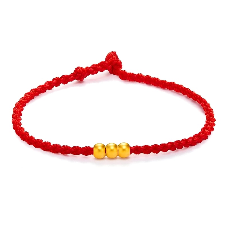 May include: A red braided bracelet with three gold beads. The bracelet is made of a tightly woven red cord, and the gold beads are evenly spaced in the center. The bracelet is designed to be worn on the wrist.