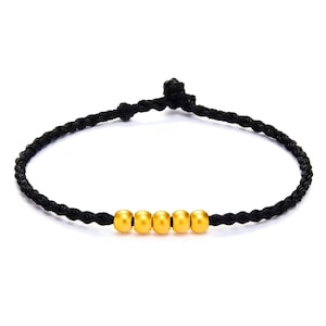 May include: A black braided bracelet featuring five gold-colored beads. The bracelet is knotted at the ends and is set against a white background. The beads are evenly spaced along the bracelet.