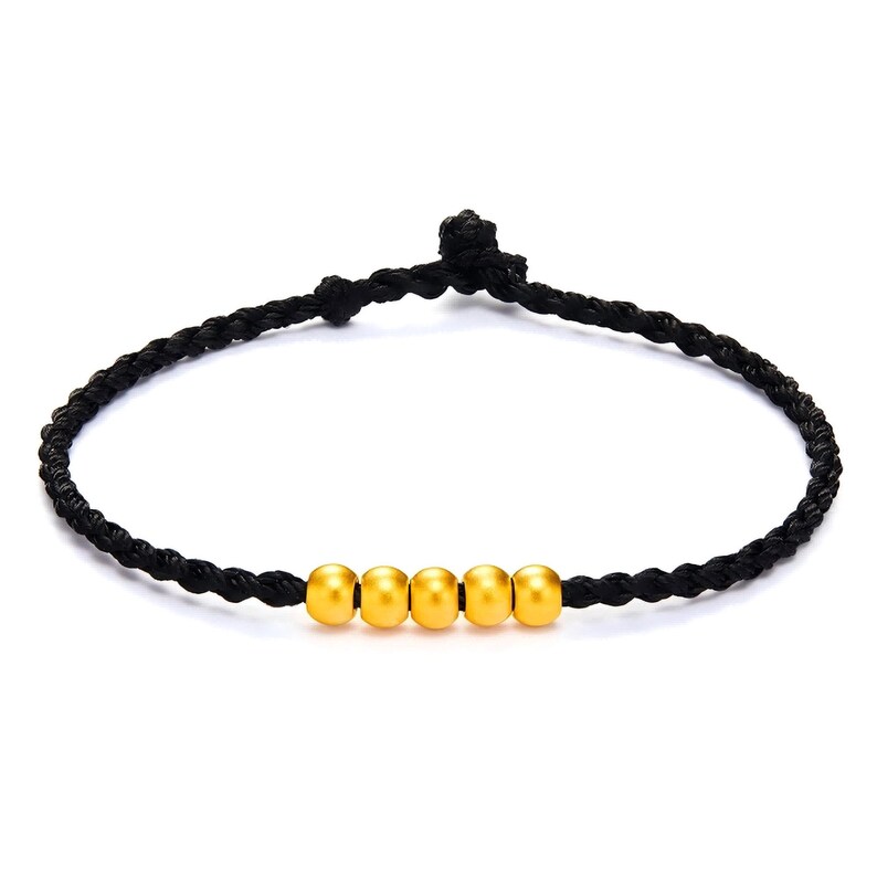 May include: A black braided bracelet featuring five gold-colored beads. The bracelet is knotted at the ends and is set against a white background. The beads are evenly spaced along the bracelet.