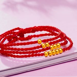 May include: Close-up of several red braided bracelets with gold-colored beads. The bracelets are layered and appear to be made of a woven material. The background is a soft pink color.