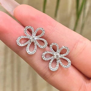 Puede incluir: Un par de pendientes de bot&oacute;n con forma de flor. Cada pendiente presenta un dise&ntilde;o floral con cinco p&eacute;talos curvos, incrustados con peque&ntilde;as piedras preciosas brillantes. El centro de cada flor tambi&eacute;n est&aacute; adornado con una piedra preciosa.