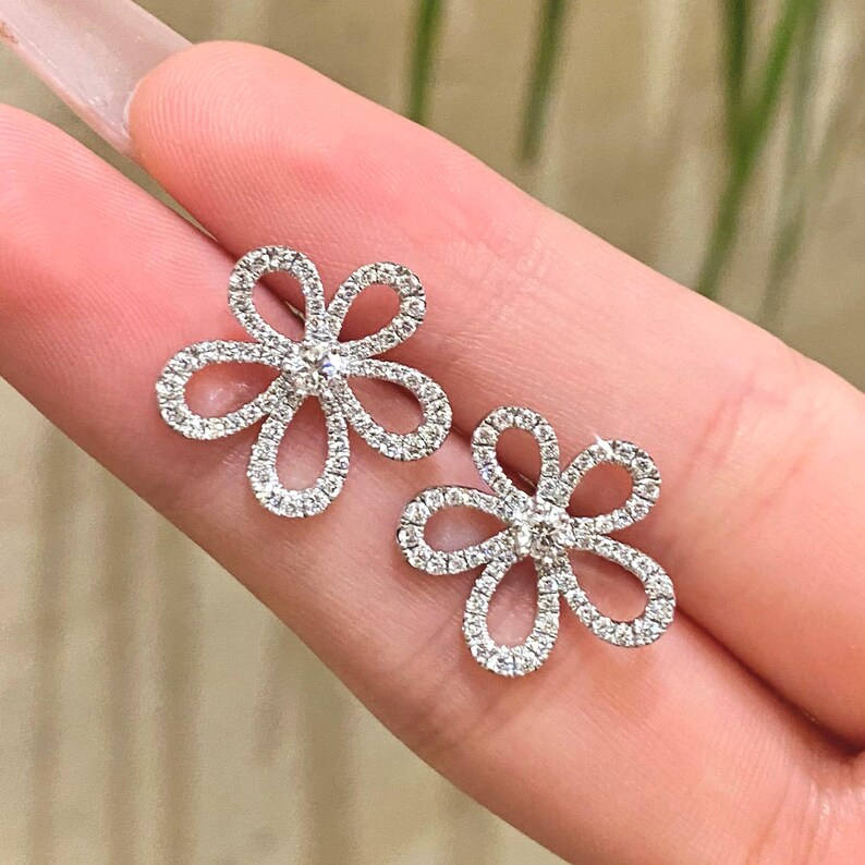 Puede incluir: Un par de pendientes de bot&oacute;n con forma de flor. Cada pendiente presenta un dise&ntilde;o floral con cinco p&eacute;talos curvos, incrustados con peque&ntilde;as piedras preciosas brillantes. El centro de cada flor tambi&eacute;n est&aacute; adornado con una piedra preciosa.