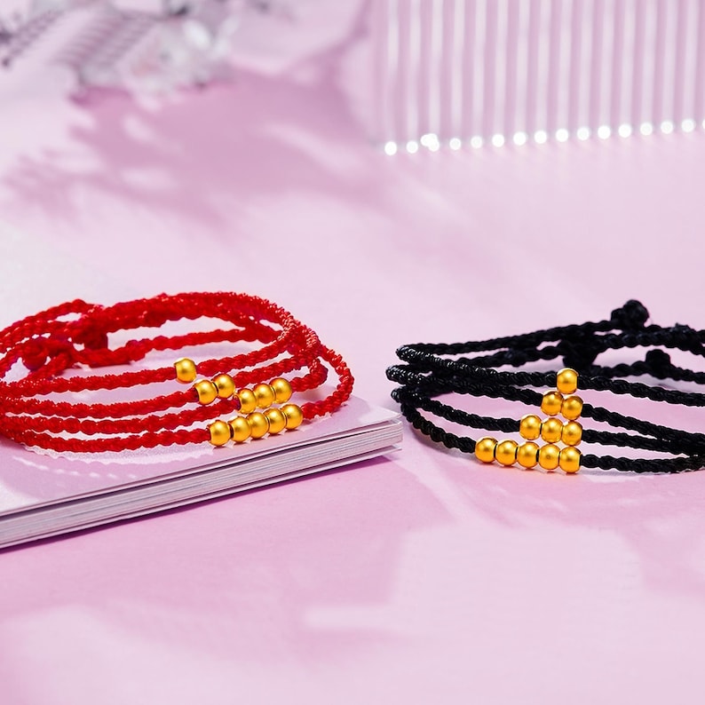 May include: Two bracelets, one red and one black, each with multiple strands and gold-colored beads. The red bracelet is on a white surface, while the black bracelet is on a pink background. The bracelets are likely for wrist wear.