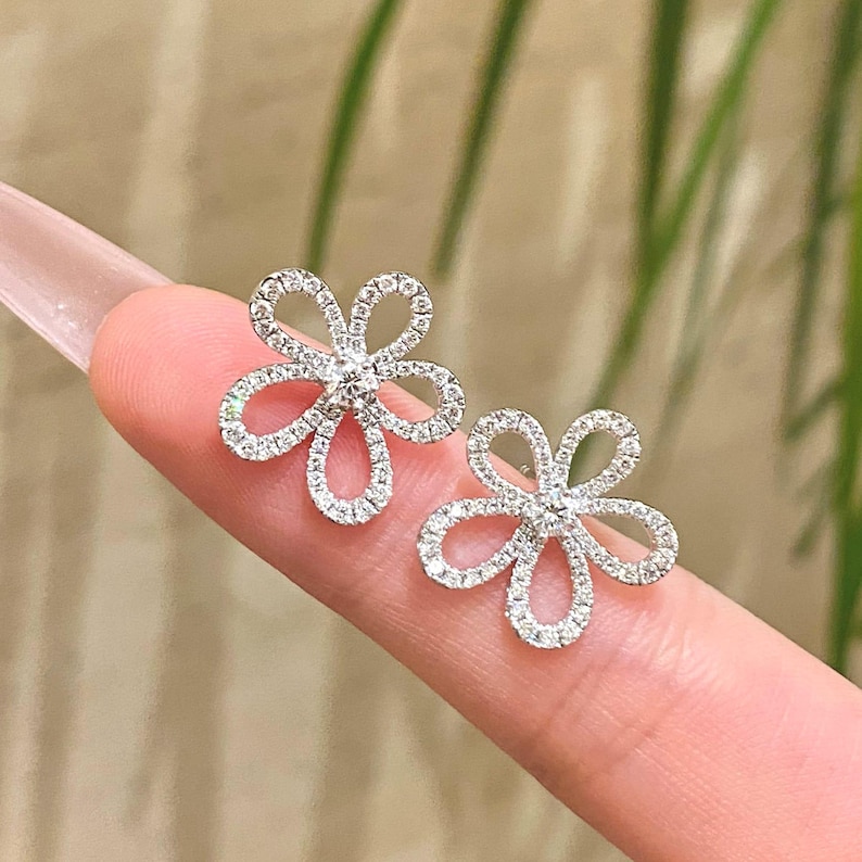 Puede incluir: Un par de pendientes de bot&oacute;n con forma de flor. Cada pendiente presenta un delicado dise&ntilde;o floral con cinco p&eacute;talos, cada uno delineado con peque&ntilde;as piedras brillantes. Los pendientes est&aacute;n sobre un fondo claro.