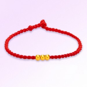 May include: A red braided bracelet with three gold beads. The bracelet is knotted at the top and has a simple, elegant design. The gold beads are evenly spaced along the bracelet's center.