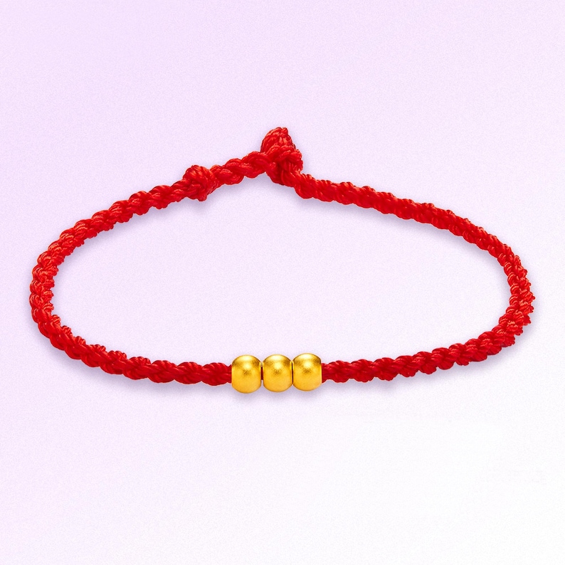 May include: A red braided bracelet with three gold beads. The bracelet is knotted at the top and has a simple, elegant design. The gold beads are evenly spaced along the bracelet's center.