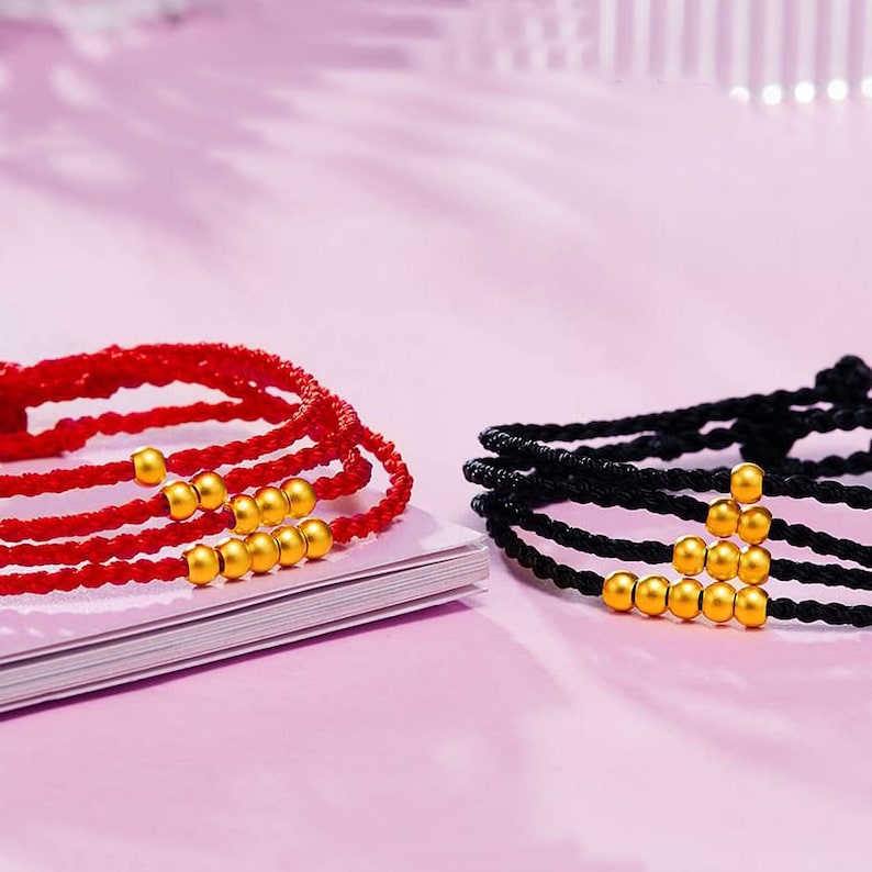 May include: Close-up of two bracelets. One is red, the other is black. Both have multiple strands of woven cord and are adorned with gold-colored beads. The bracelets are resting on a white surface against a pink background.