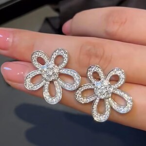 Puede incluir: Un par de pendientes de bot&oacute;n plateados con forma de flor. Cada pendiente presenta cinco p&eacute;talos curvos incrustados con peque&ntilde;as piedras preciosas transparentes. El centro de cada flor es un grupo de piedras preciosas transparentes m&aacute;s grandes.