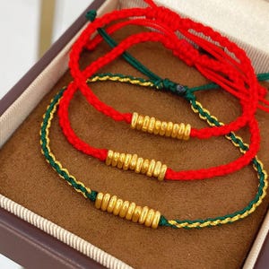 Op de afbeelding: Close-up van drie rood en groen gevlochten armbanden met goudkleurige kralen. De armbanden worden in een bruine doos getoond. De armbanden zijn gemaakt van rood en groen gevlochten koord met goudkleurige kralen.