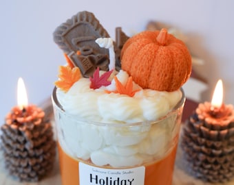 May include: A decorative candle with a pumpkin and fall leaf design. The candle is in a clear glass with orange wax and white frosting-like wax on top. The label reads "Holiday Spice". Two pinecone candles flank the main candle.