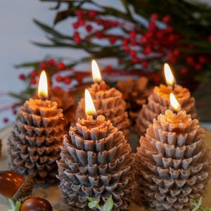 May include: Six lit pine cone-shaped candles with a brown hue are arranged on a wooden surface. Acorns and greenery are scattered around the candles. Red berries and green leaves are in the background.