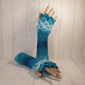 May include: A pair of blue and teal crochet fingerless gloves with a scalloped design. The gloves are made with a gradient yarn that transitions from a light blue to a darker teal.