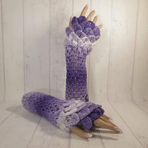 May include: A pair of long, crocheted fingerless gloves with a gradient of purple and white. The gloves have a scalloped edge and a delicate, lacy pattern.