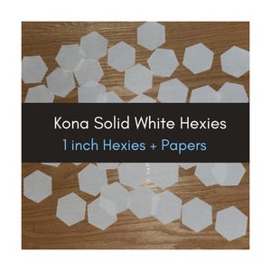 May include: White hexagonal paper shapes scattered on a wooden surface. The image includes text that reads "Kona Solid White Hexies" and "1 inch Hexies + Papers". These shapes are likely for crafting or quilting.