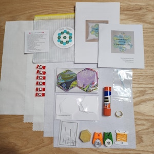 May include: A craft kit with various sewing supplies, including fabric, paper templates, fabric clips, a glue stick, and thread. The kit also includes two hexagon-shaped fabric pieces with floral patterns and a beginner's guide.