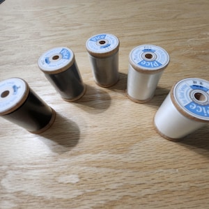 May include: Five spools of thread in various colors, including black, gray, and white, are arranged on a wooden surface. Each spool has a wooden base and a paper label with text. The spools are for sewing and crafting.