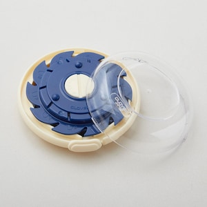 May include: A round Clover brand sewing tool with a cream-colored outer ring and a dark blue rotating inner disc featuring numbered notches. A clear, circular plastic lid is detached and resting beside the tool, revealing the Clover logo.