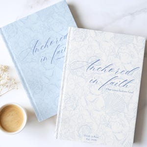 May include: Two hardcover journals with floral patterns. One is light blue, the other is white. Both have the text "Anchored in faith" in cursive. The white journal has the text "Our vows before God" and "Emily & John Est. 2020".