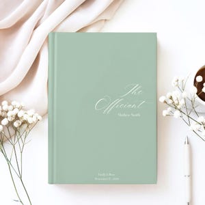 Puede incluir: Un libro verde salvia con el t&iacute;tulo "The Officiant" en escritura cursiva. El nombre Mathew Smith est&aacute; debajo. El libro est&aacute; rodeado de flores blancas, un bol&iacute;grafo y una taza de caf&eacute;.