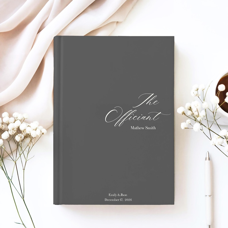 Puede incluir: Un libro gris oscuro con las palabras "The Officiant" y el nombre Mathew Smith en elegante escritura blanca. Los nombres Emily & Jhon y la fecha 17 de diciembre de 2026 est&aacute;n impresos en la parte inferior. El libro est&aacute; rodeado de flores blancas y un bol&iacute;grafo.