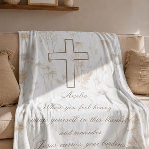 Puede incluir: Una manta blanca con un gran diseño de cruz y detalles florales. La manta tiene el texto: "Amelia, Cuando te sientas pesado, envuélvete en esta manta y recuerda: Jesús lleva tus cargas."