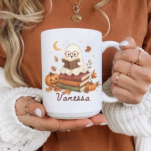 May include: A white ceramic mug featuring a graphic of a ghost wearing round glasses and reading a book while sitting on a stack of three books. The design includes two pumpkins, autumn leaves, a crescent moon, and the name Vanessa in script.
