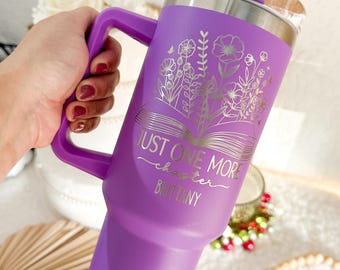 May include: A purple stainless steel tumbler with a handle and a clear lid featuring a purple top. The tumbler displays a gold-colored line art graphic of an open book with flowers growing from the pages, the text JUST ONE MORE chapter, and the name BRITTANY.