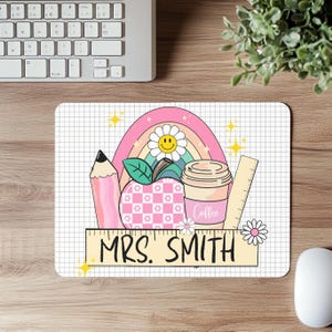 Può includere: Un tappetino per mouse personalizzato con una matita rosa, una mela, una tazza di caffè e un righello. Sono presenti anche un arcobaleno e una margherita sorridente. Il tappetino ha il testo "MRS. SMITH" su uno striscione giallo.