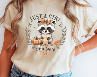 Op de afbeelding: Beige T-shirt met een illustratie van een wasbeer die een koffiekopje, een pompoen en herfstbladeren vasthoudt. De tekst luidt "Just a girl who loves FALL". De wasbeer heeft een strik. Het shirt is neutraal van kleur.