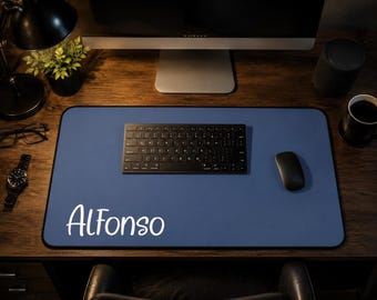 May include: A rectangular blue desk mat with a black border and the name Alfonso printed in white script lettering. The mat is positioned on a dark wood surface, serving as a base for a black wireless keyboard and a black computer mouse.
