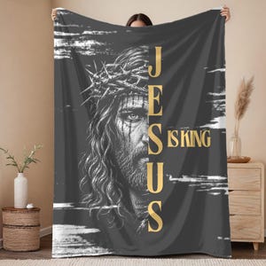 Könnte beinhalten: Eine dunkelgraue Wohndecke mit einer schwarz-weißen Illustration eines Gesichts mit Dornenkrone. Der Text "JESUS IS KING" ist in vertikaler goldener Schrift auf der rechten Seite aufgedruckt, ergänzt durch weiße horizontale Pinselstriche.
