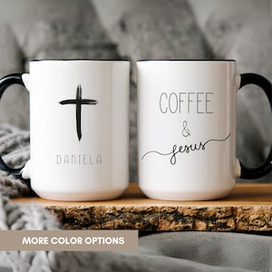 May include: Two white ceramic mugs with black handles. The left mug features a black brushstroke cross above the name DANIELA in a sans-serif font. The right mug displays the text COFFEE & Jesus in a mix of block and cursive lettering.