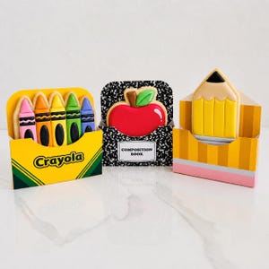 Puede incluir: Una colección de galletas con temática de regreso a clases. Las galletas incluyen una caja de crayones, un cuaderno de composición con una manzana y un lápiz. La caja de crayones es amarilla con el logo de Crayola. La galleta de manzana es roja con una hoja verde.