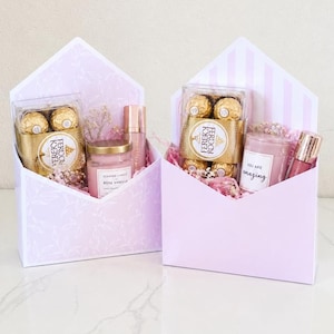 May include: Two gift boxes shaped like envelopes, filled with treats. Each box contains Ferrero Rocher chocolates, a scented candle, and a beauty product. The boxes are pink with floral and striped patterns, and are filled with pink filler.