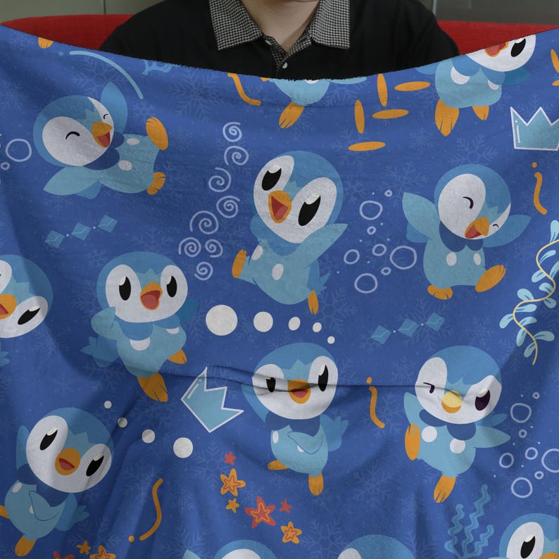 May include: A blue fleece blanket featuring a repeating pattern of light blue and white penguin characters with orange beaks and feet. The design includes scattered white snowflakes, yellow stars, blue crowns, and small orange and yellow shapes.