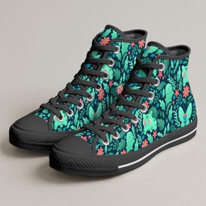 Puede incluir: Zapatillas altas de lona con un fondo verde oscuro y un patrón repetido de criaturas de dibujos animados verdes, flores y hojas. Las zapatillas tienen cordones negros, punteras negras y una entresuela blanca con una raya negra.