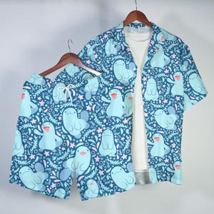 以下が含まれることがあります： ボタンダウンシャツとショートパンツを含む、お揃いの服のセット。ダークティール色の背景に、笑顔の漫画の生き物、星、葉のティールパターンが特徴です。 シャツは開いており、白いアンダーシャツが見えます。 ショーツにはドローストリングが付いています。