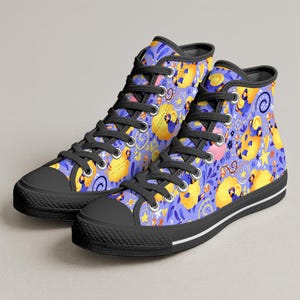 Könnte beinhalten: Hohe Canvas-Sneaker mit einem skurrilen Muster. Die Schuhe haben schwarze Zehenkappen, Schnürsenkel und Sohlen. Der gemusterte Stoff ist hellviolett mit gelben, rosa und blauen Cartoon-Illustrationen. Ein schwarzer Streifen verläuft entlang der Sohle.