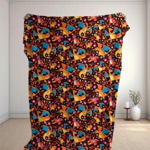 Puede incluir: Una manta rectangular con un patrón repetitivo de criaturas naranjas parecidas a dragones con alas azules, pequeñas criaturas rosas y esferas rojas y blancas sobre un fondo marrón oscuro. El diseño incluye pequeñas estrellas blancas y llamas.